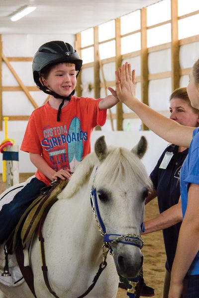 A young boy high-fives volunteers while riding a therapeutic riding horse